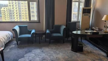 A hotel room with a bed, two armchairs, a small table, a desk with a lamp, and a large window with a city view.