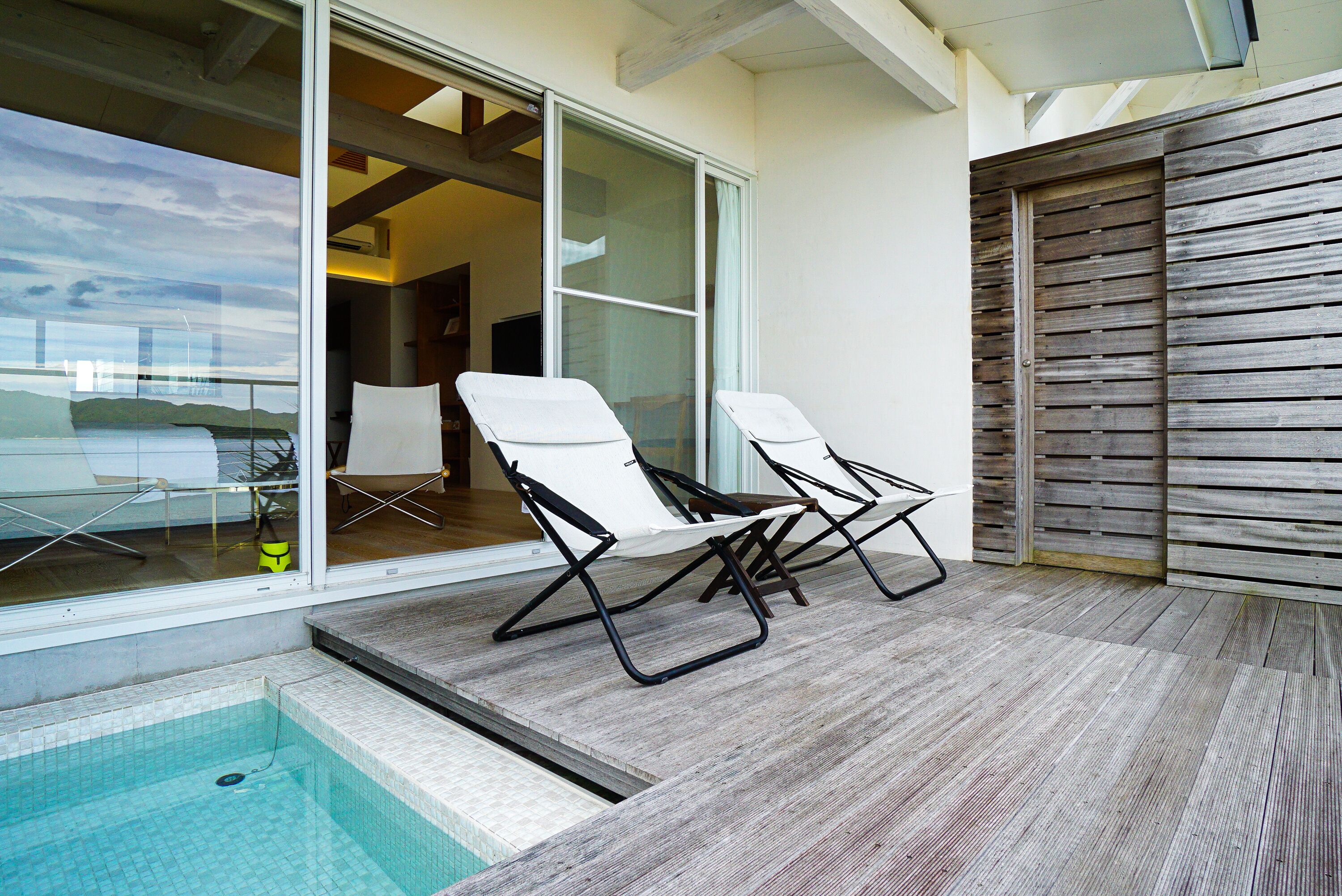 Area piscina moderna all'aperto con due lettini, una porta in vetro che conduce alla piscina e un muro in legno.