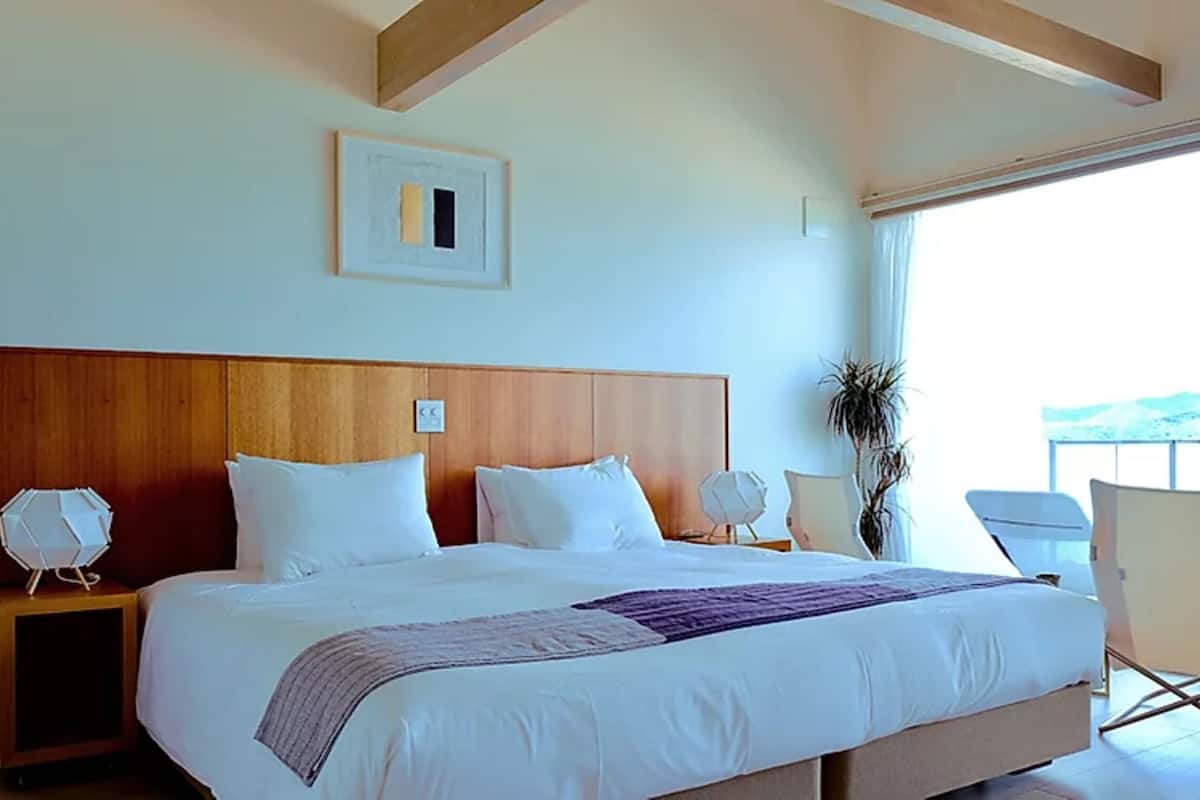 A hotel room with a large bed, a wooden headboard, a bedside table with a lamp, a chair, and a view through the window.