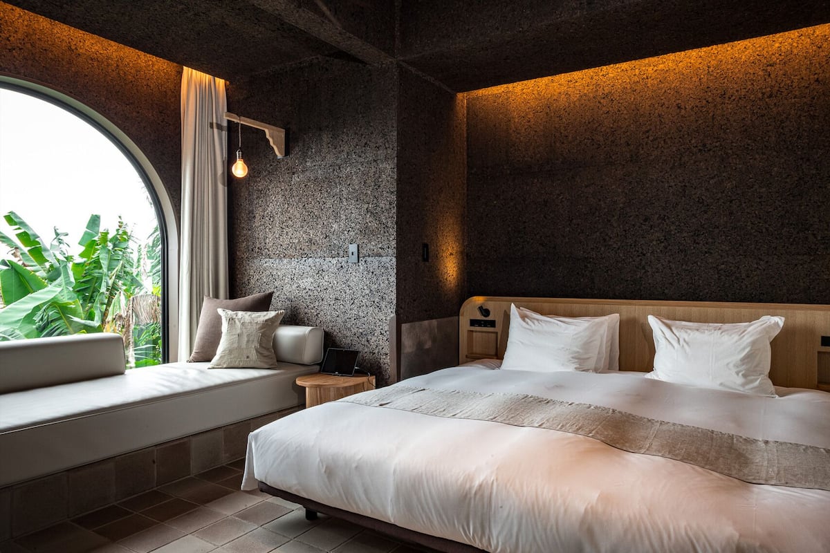 A modern bedroom with a large bed, a bedside table, a nightstand, a window with a view of greenery, and a textured wall.