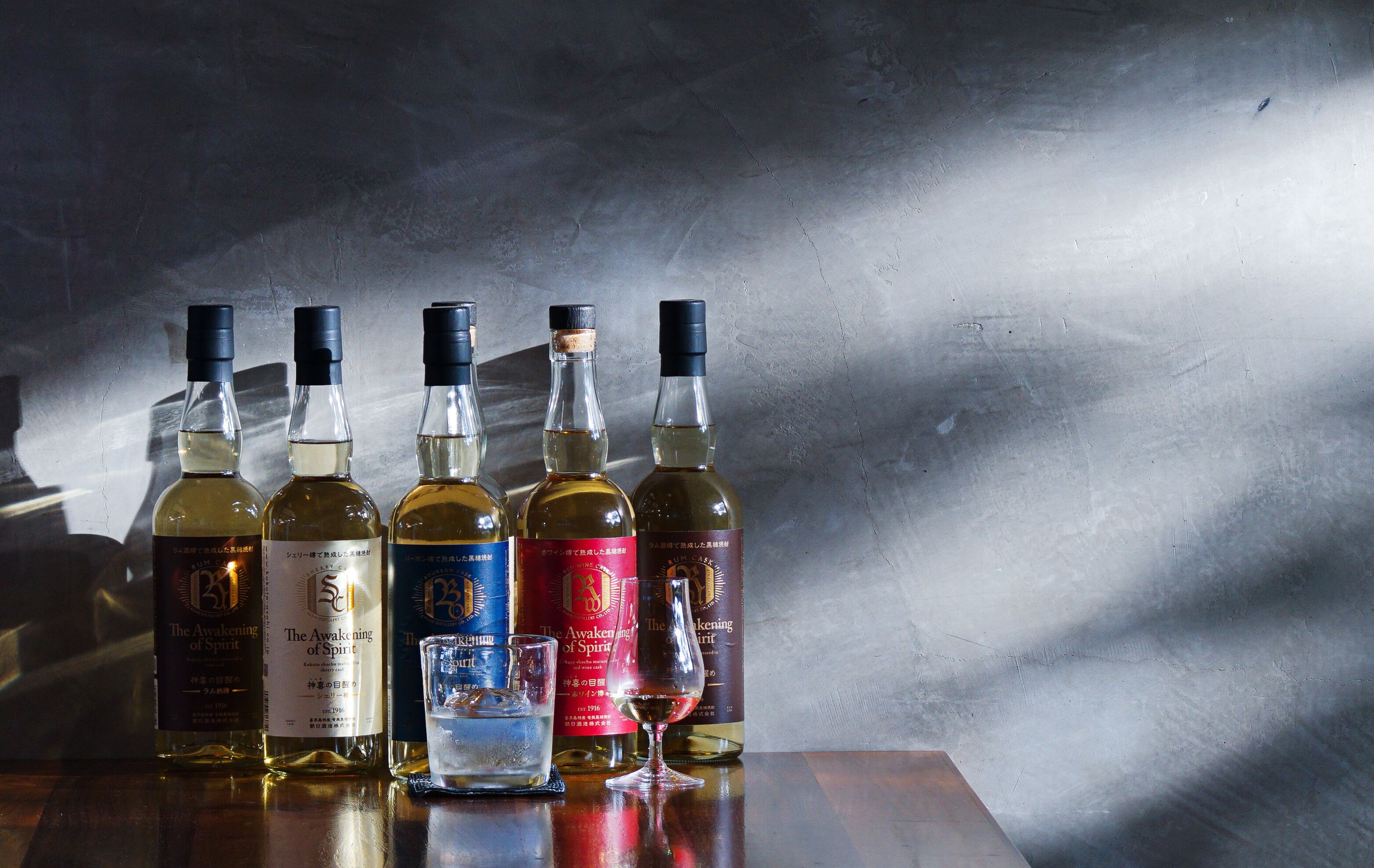 Varias botellas de whisky sobre una superficie reflectante, con una pared texturizada al fondo.