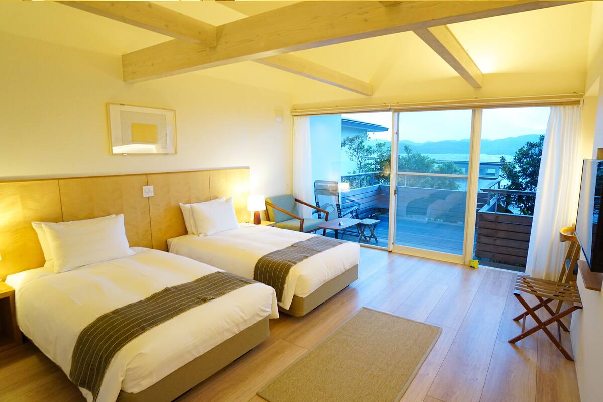 A hotel room with two beds, a large sliding glass door leading to a balcony, and a view of the outdoors.