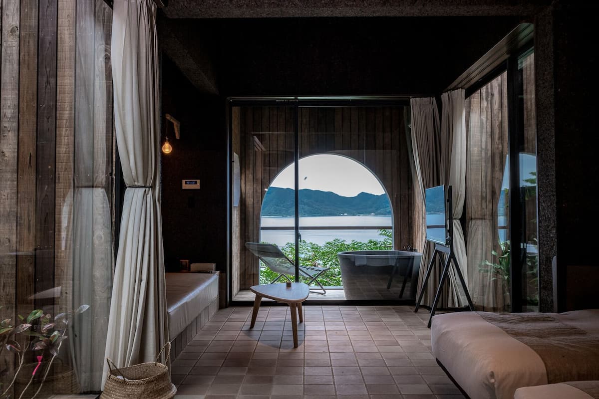 A room with a large window offering a view of a lake and mountains, a wooden bench, a chair, and a bed.