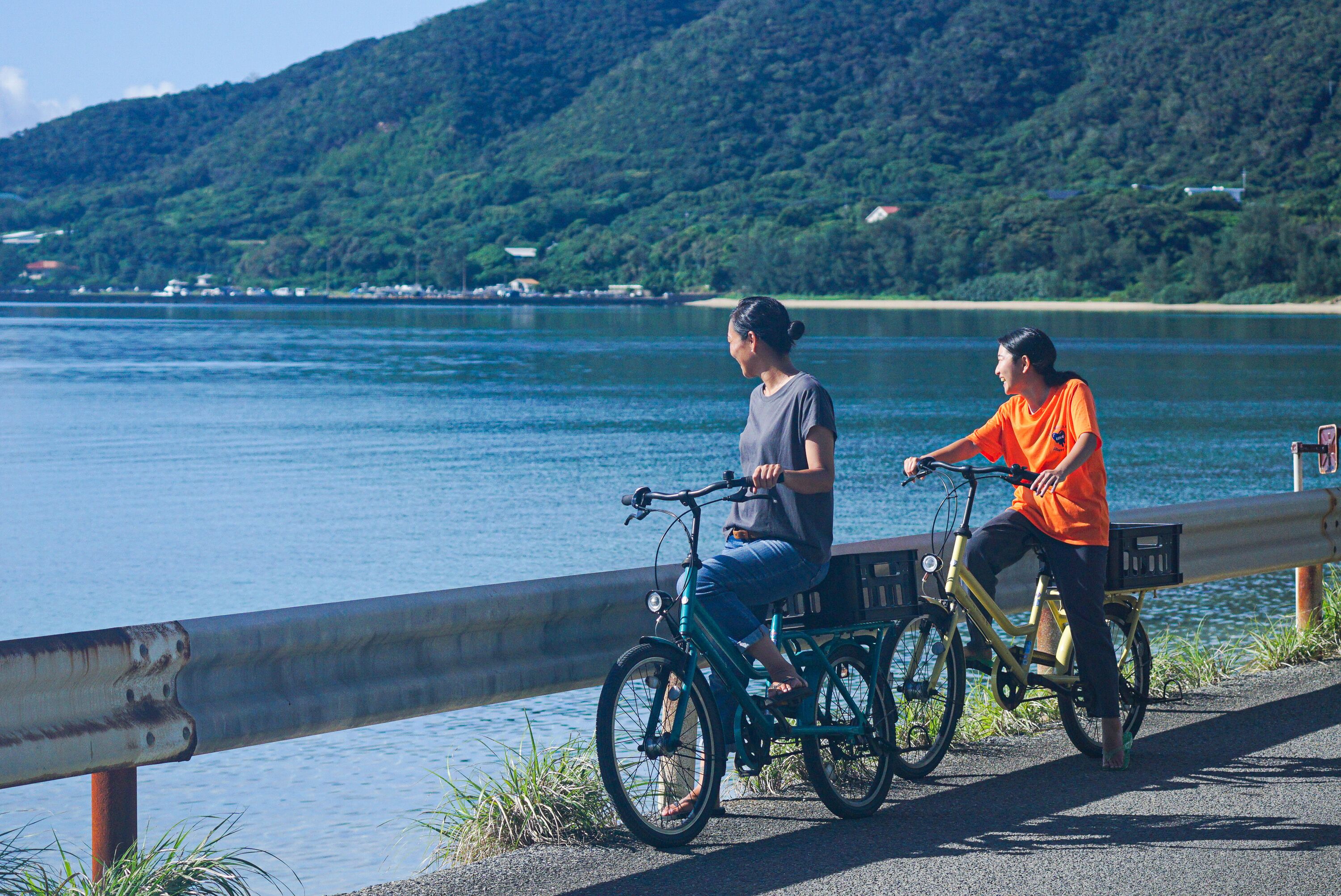 Two people riding bicycles along a waterfront path with a metal railing and a view of a lake and hills in the background.