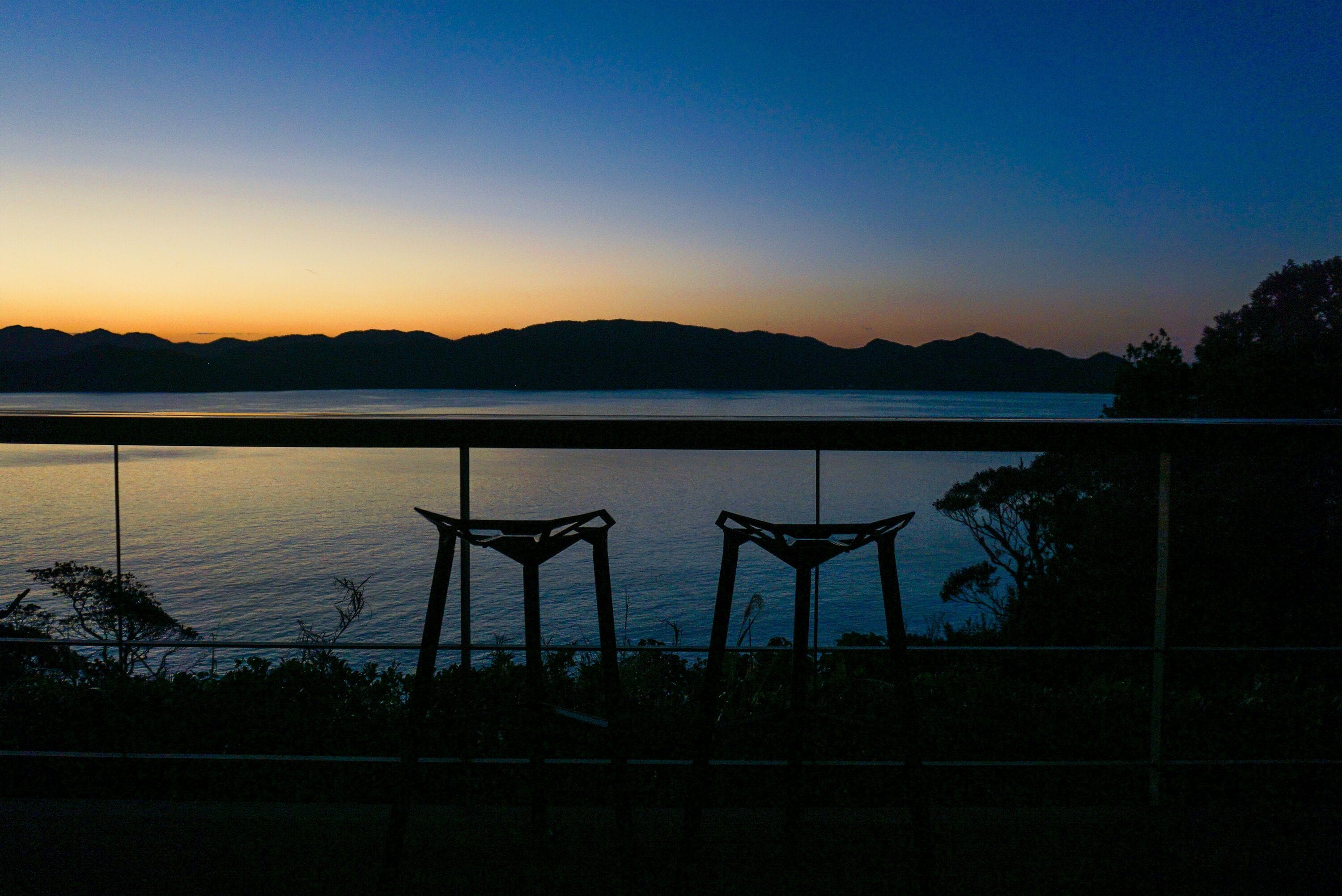A sunset view from a balcony with two chairs facing a calm body of water and distant mountains.