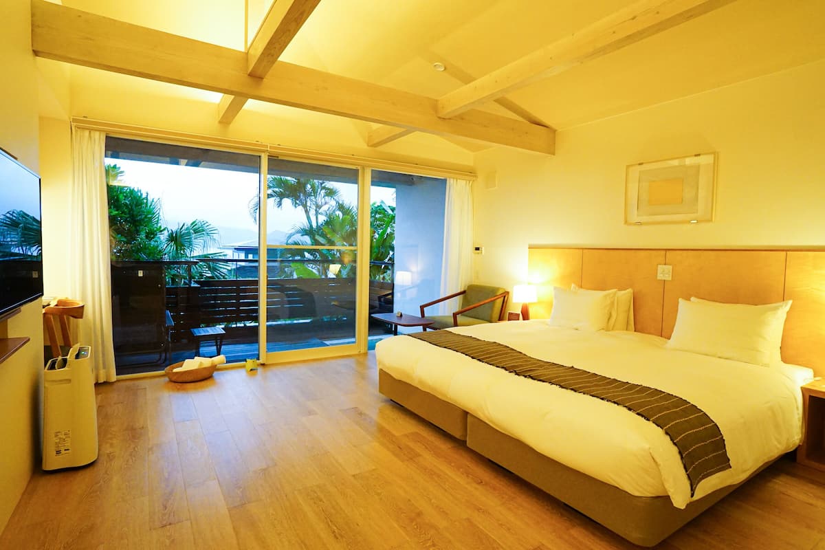 A hotel room with a large bed, a TV, a desk, and a balcony with a view of palm trees.