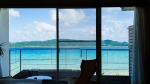 A room with a large sliding glass door leading to a balcony with a view of the sea and mountains. - Miru Amami (Tatsugo)