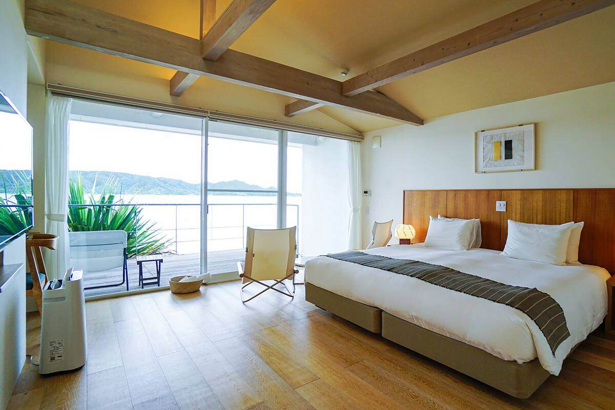 A bedroom with a large bed, a window with a view, a chair, and a potted plant.