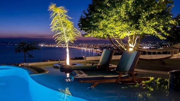 An infinity pool with a view of the city at night, surrounded by lounge chairs and a hammock. - Villa Paradizo (Kalamata)