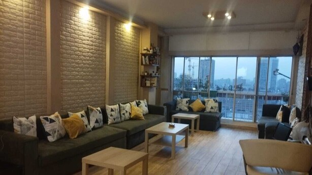 Un salon moderne avec un canapé d’angle, deux tables basses et une vue sur la ville grâce à de grandes fenêtres.