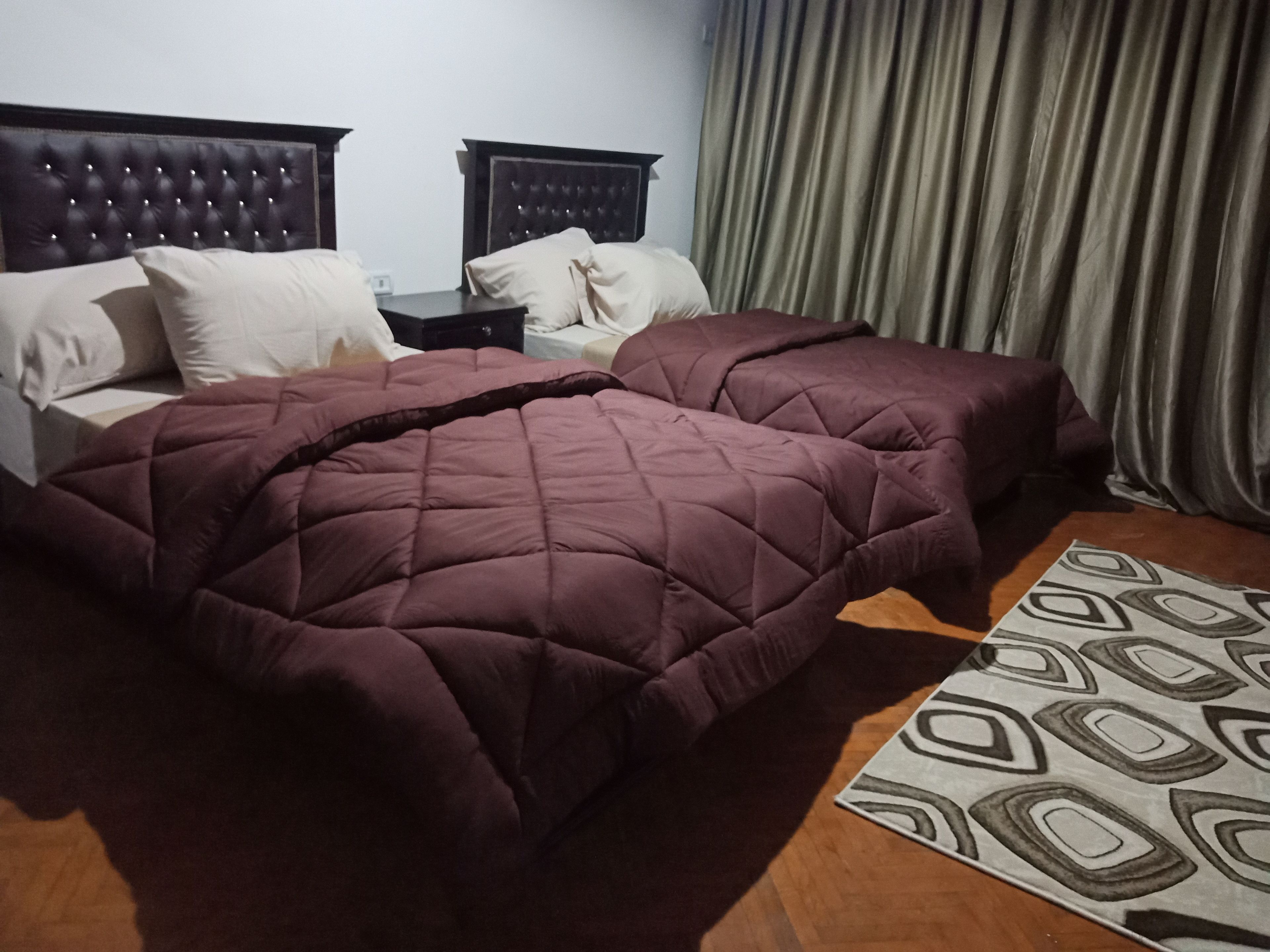 A hotel room with two beds, a wooden floor, a patterned rug, and a curtain.