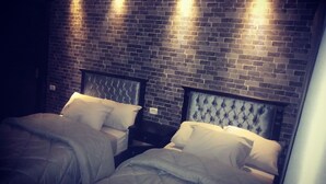 Une chambre avec deux lits, chacun doté d’une tête de lit et de linge de lit blanc. Les murs sont en briques sombres, et trois lampes sont fixées au mur.
