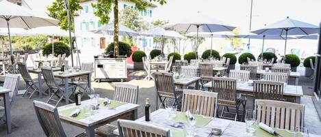 Un espace extérieur aménagé pour les repas, avec des tables, des chaises, des parasols et un bâtiment en arrière-plan.