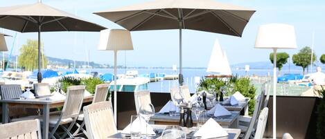 Un área de comedor al aire libre con mesas, sillas y sombrillas con vista a un puerto deportivo y barcos.