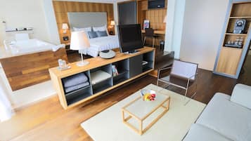 A modern hotel room with a wooden floor, a bed with a headboard, a TV on a wooden stand, a small table with a bowl of fruit, and a sofa.