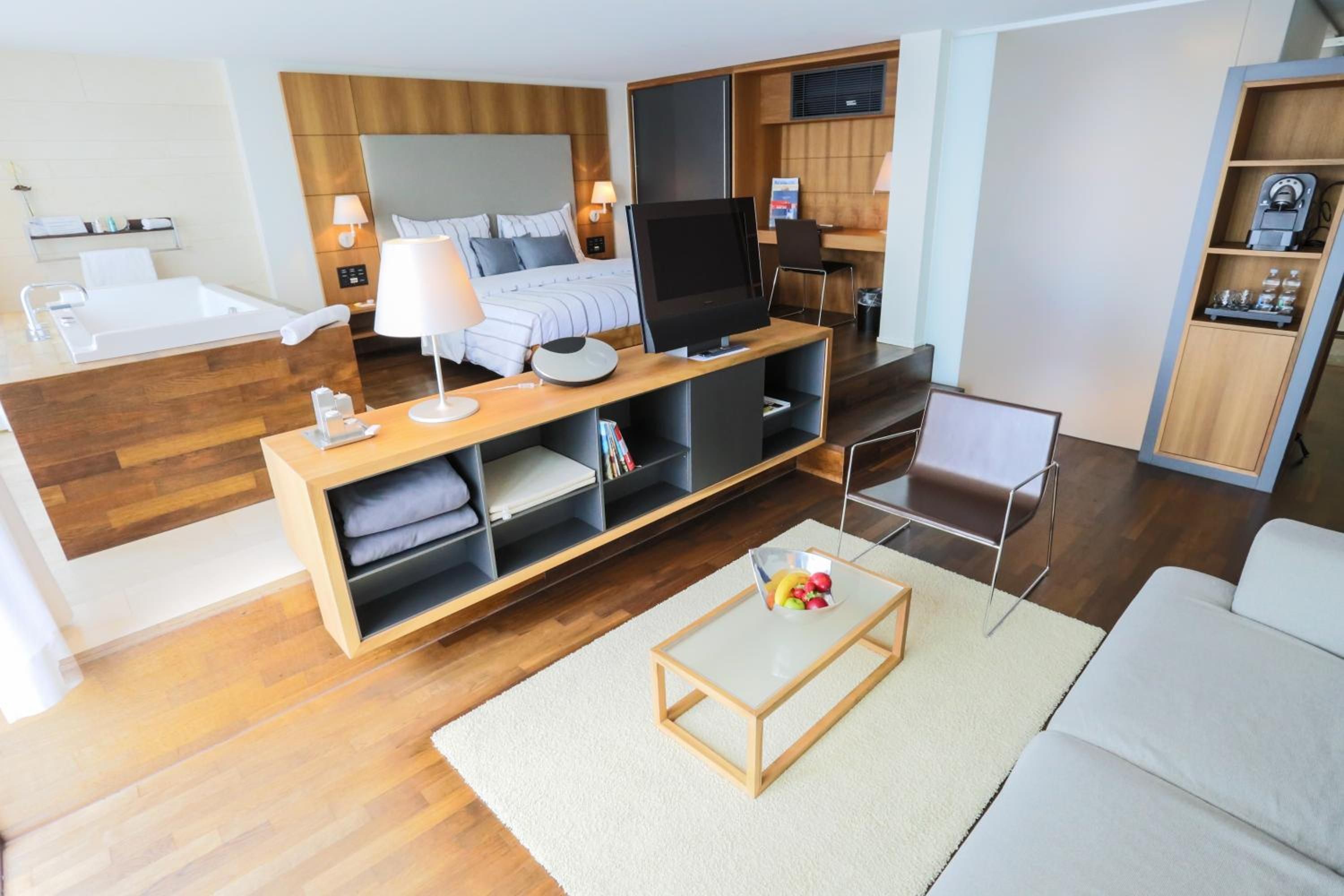 Habitació d'hotel moderna amb sòl de fusta, un llit amb capçalera, un televisor sobre un suport de fusta, una taula petita amb un bol de fruita i un sofà.