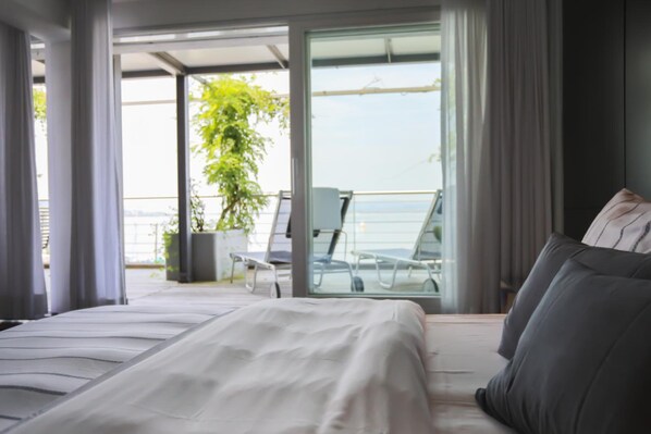 A modern bedroom with a large bed, a balcony with a view of the sea, and a potted plant.