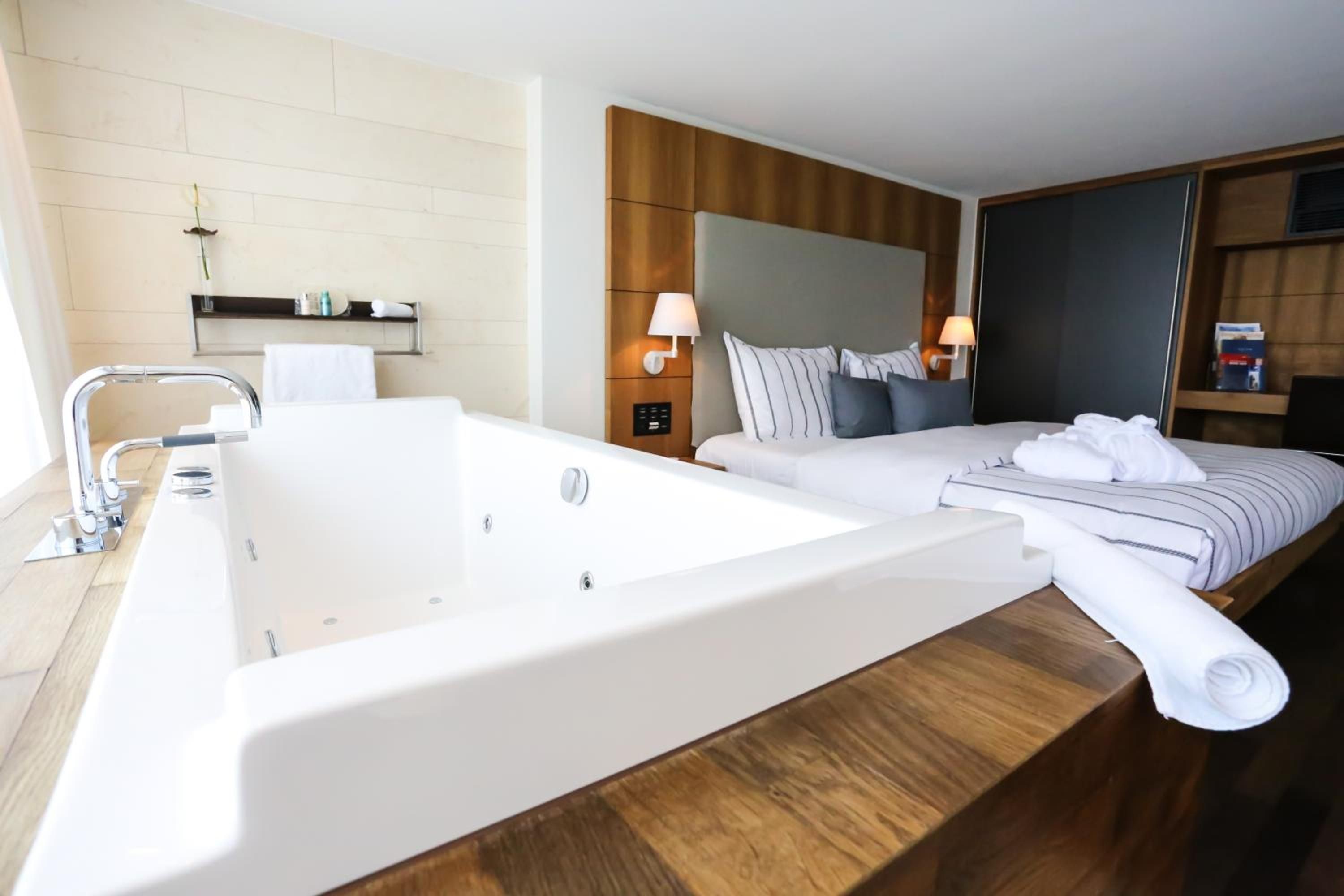 Habitación de hotel con una bañera grande, cama con almohadas, mesita de noche y televisor instalado en la pared.