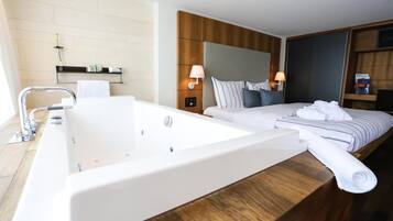 Habitación de hotel con una bañera grande, cama con almohadas, mesita de noche y televisor instalado en la pared.