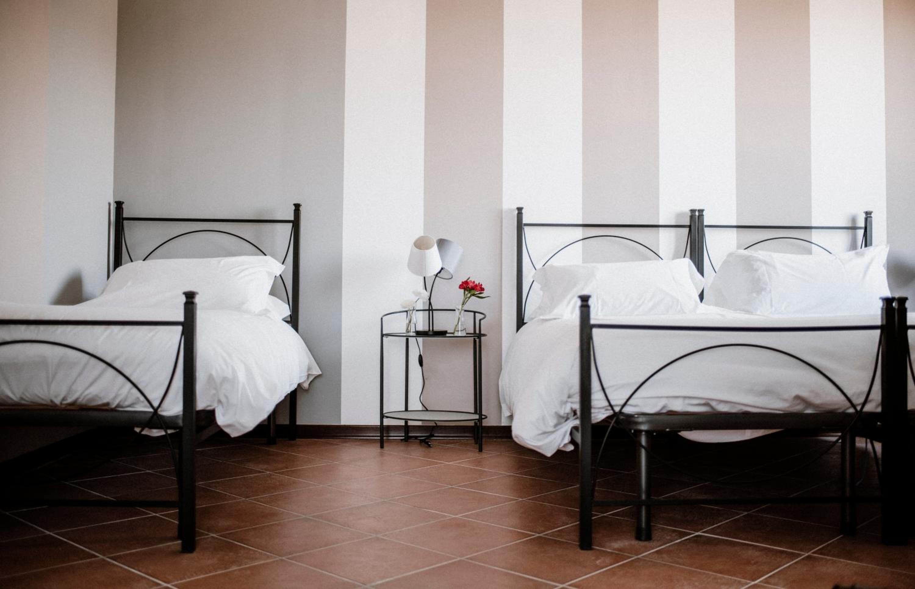 Duas camas de solteiro com estrutura de metal, roupa de cama branca e uma pequena mesa de cabeceira com abajur e um vaso de flores.