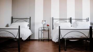 Duas camas de solteiro com estrutura de metal, roupa de cama branca e uma pequena mesa de cabeceira com abajur e um vaso de flores.