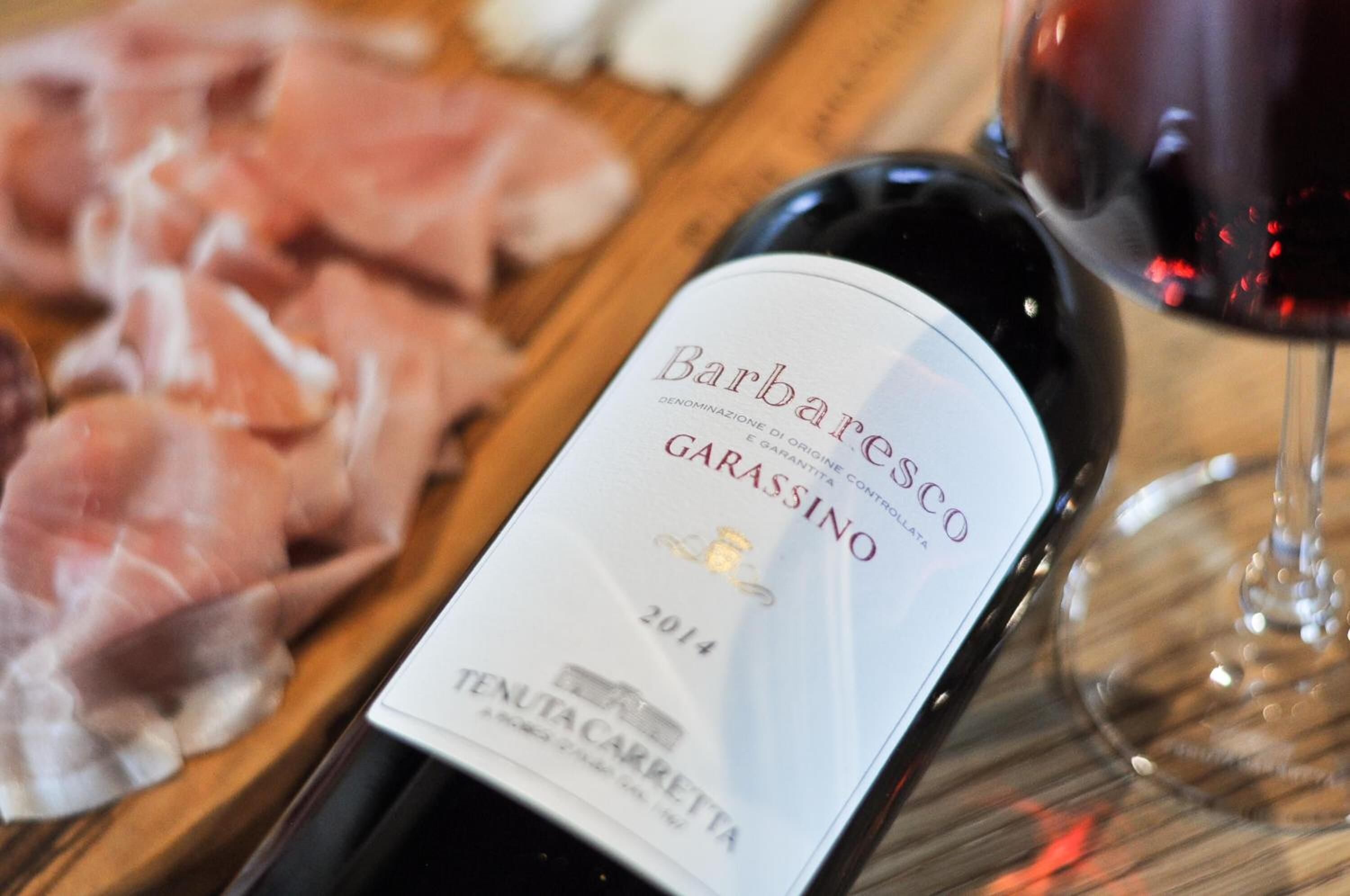 A bottle of Barbaresco Garassino wine from 2014, a wooden board with sliced meats, and a glass of red wine.