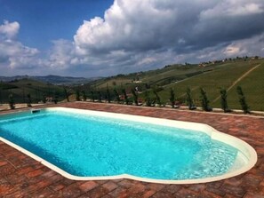 Una piscina rectangular con solárium empedrado, rodeada por un paisaje montañoso con vegetación escasa y edificios a lo lejos.