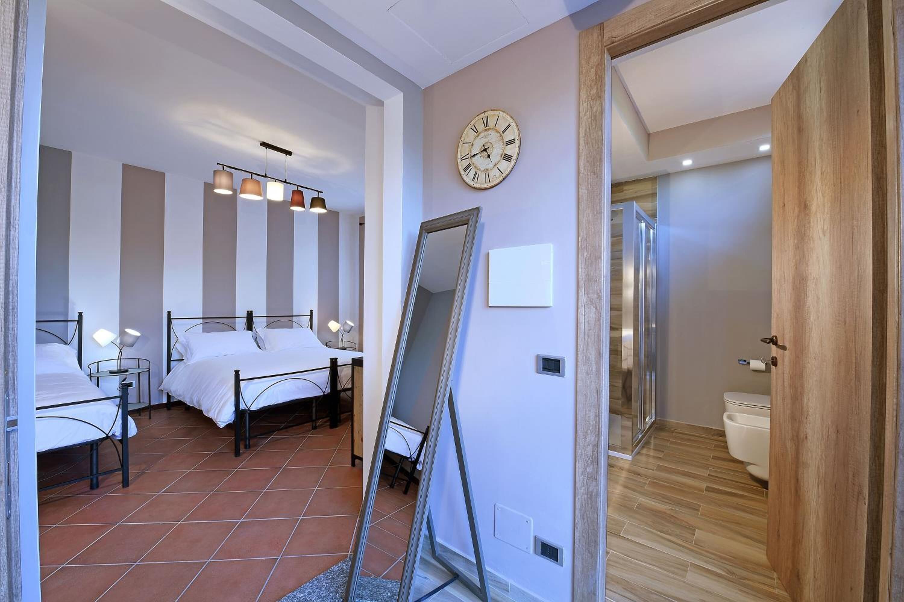 Habitación de hotel con dos camas, un espejo grande, un reloj en la pared y una puerta de madera que da al baño.