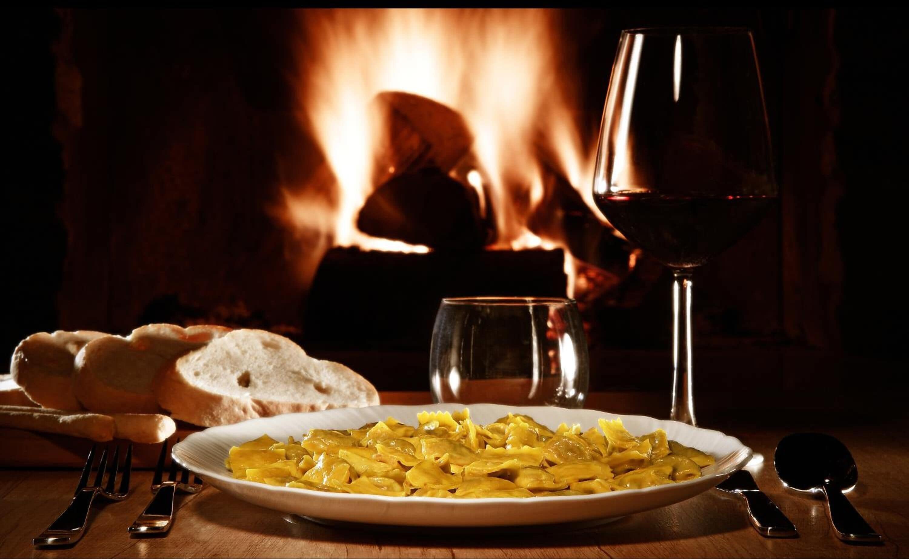 Un plato de pasta, una copa de vino tinto, pan y cubiertos sobre una mesa con una chimenea al fondo.