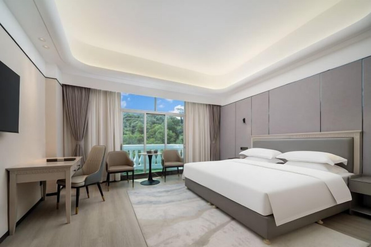 A modern hotel room with a large bed, a desk, two chairs, and a balcony with a view of greenery.