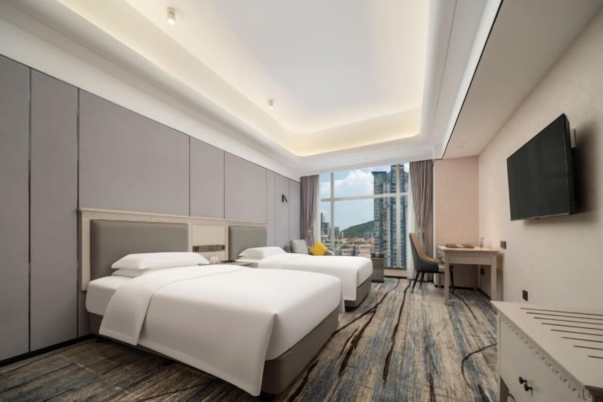 A hotel room with two beds, a desk, a chair, a TV, and a large window with a city view.