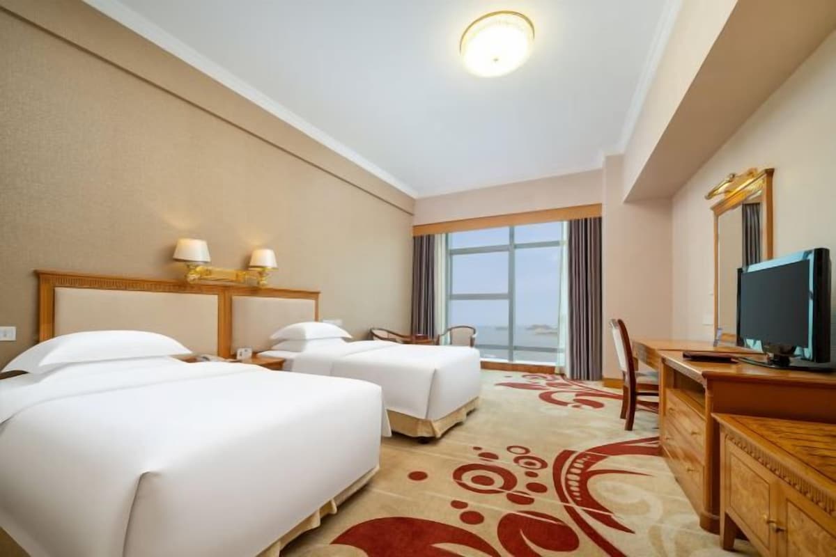 A hotel room with two beds, a desk, a television, and a large window.
