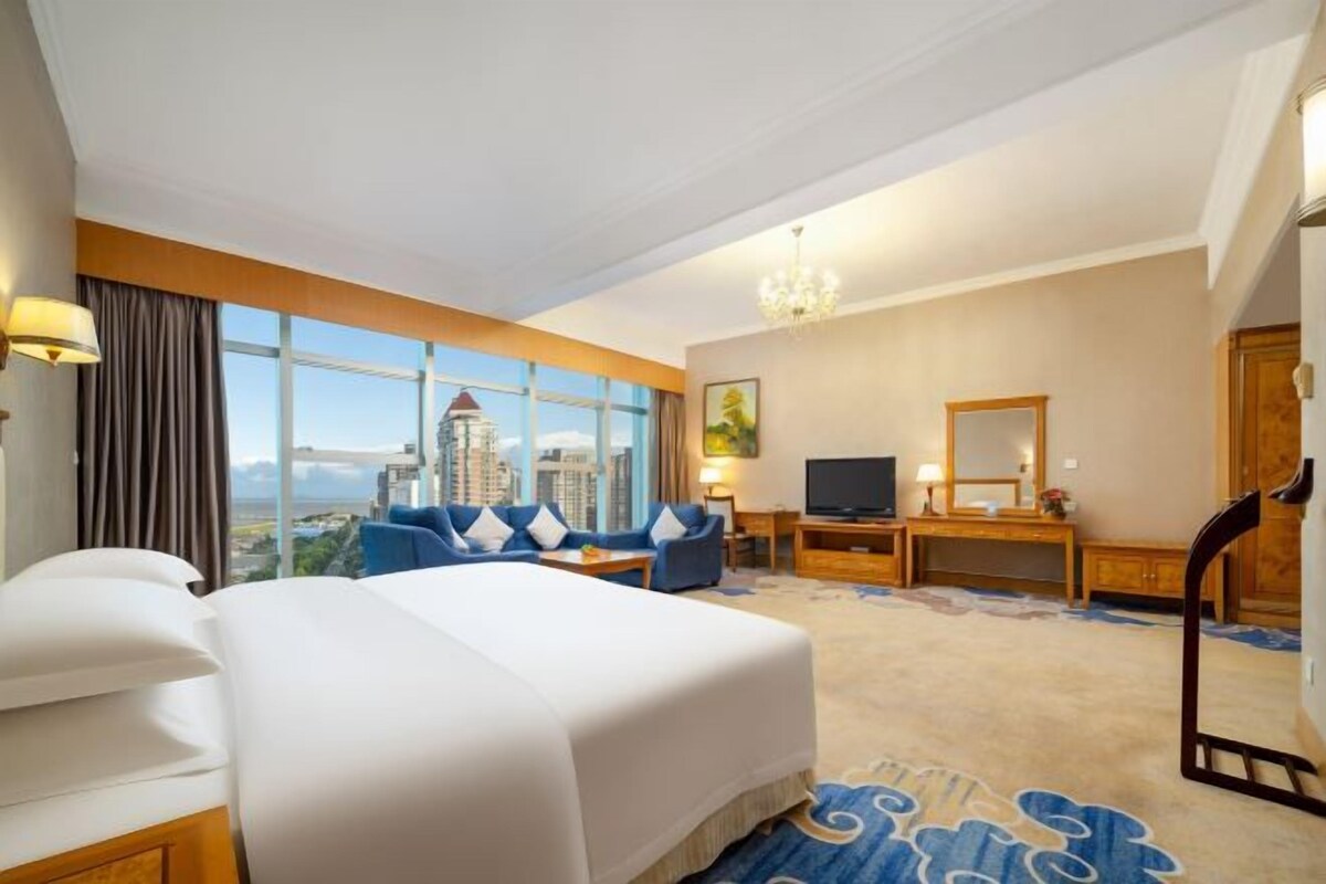 A hotel room with a large bed, a sofa, a desk with a TV, and a view of the cityscape.