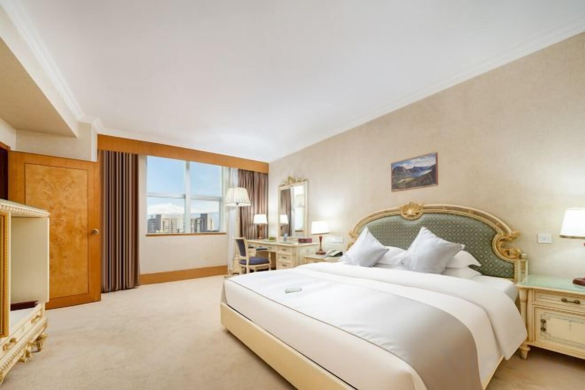 A hotel room with a large bed, a desk, a chair, a dresser, and a window with a city view.