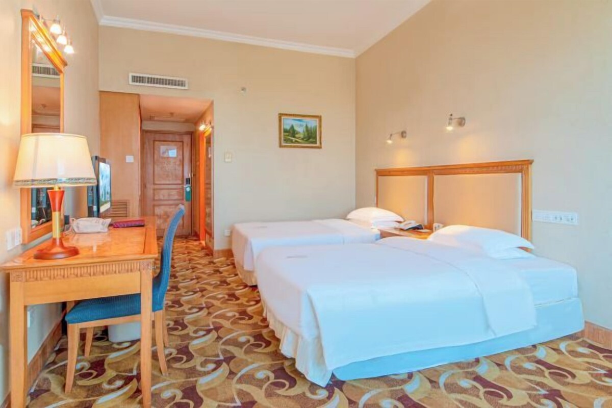 A hotel room with two single beds, a desk with a lamp, a chair, and a patterned carpet.