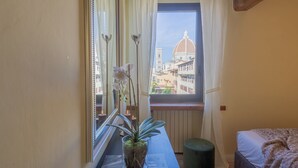 Une chambre avec une fenêtre donnant sur le paysage urbain, un lit, une table de chevet avec une plante et un pouf vert.