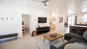 A modern living room with a flat-screen TV, a ceiling fan, a coffee table, a sofa, and a kitchen area with bar stools. - New poolside Luxury townhouse in The Lofts (St. George)
