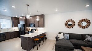 A modern kitchen with dark cabinets, a central island with stools, a refrigerator, and a microwave. There is a dark-colored sofa and a patterned rug in the living area. - New poolside Luxury townhouse in The Lofts (St. George)