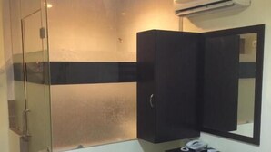 A compact room with a glass shower enclosure, a dark wooden cabinet, a small bedside table with a phone, and a wall-mounted air conditioner. - Rafayeh Hotel (Seremban)