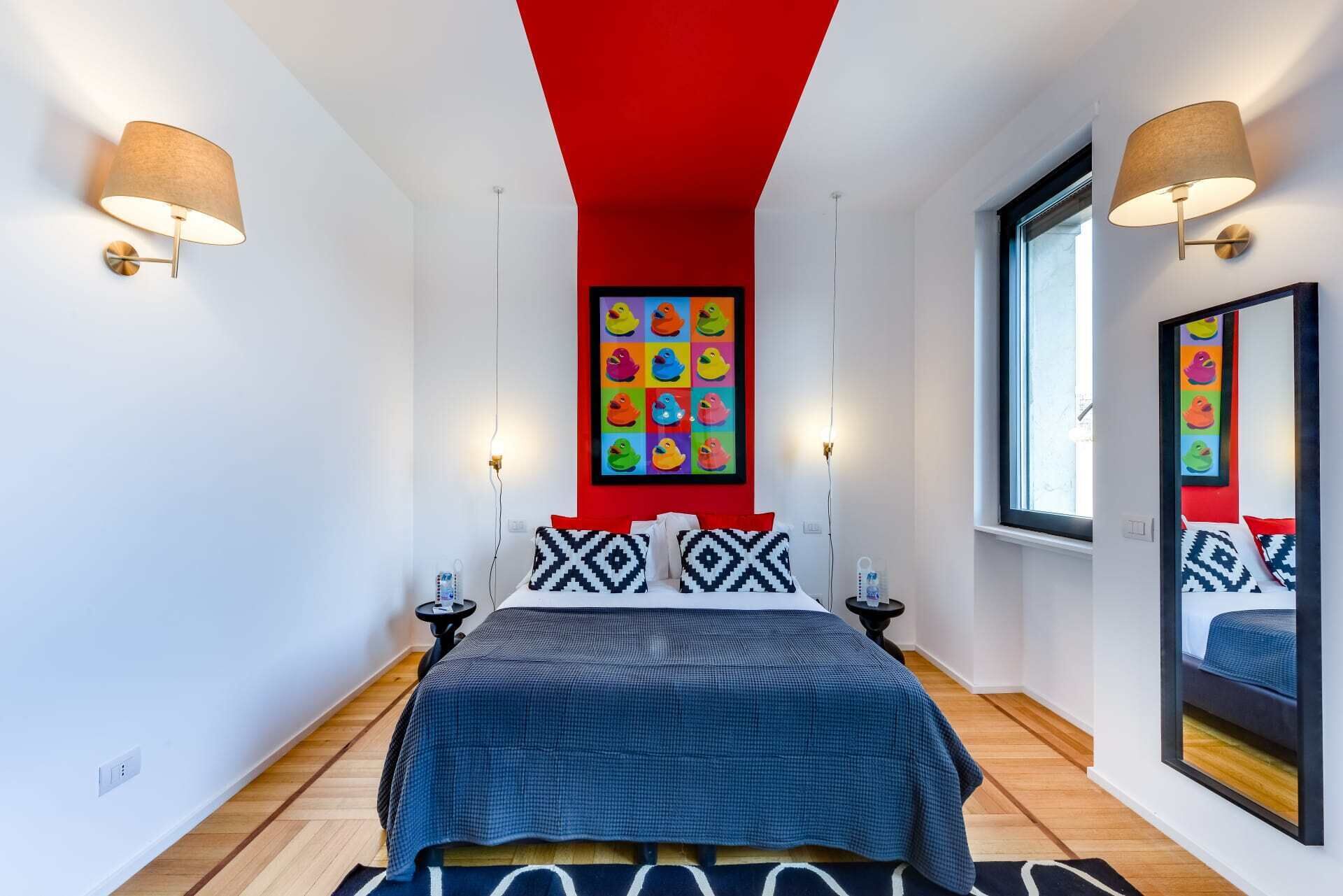 Ein modernes Schlafzimmer mit roter Decke, einem großen Bett mit gemusterten Kissen, zwei Wandleuchten und einem farbenfrohen Kunstwerk über dem Bett.