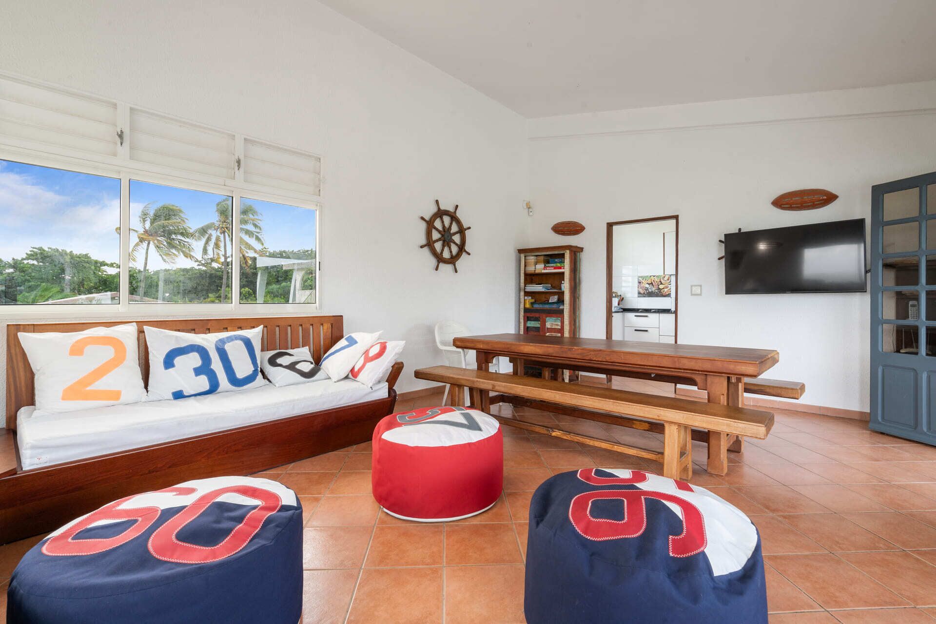 Una habitación con sofá, otomanes, una mesa de madera, una estantería, televisor y ventana con vista a palmeras.