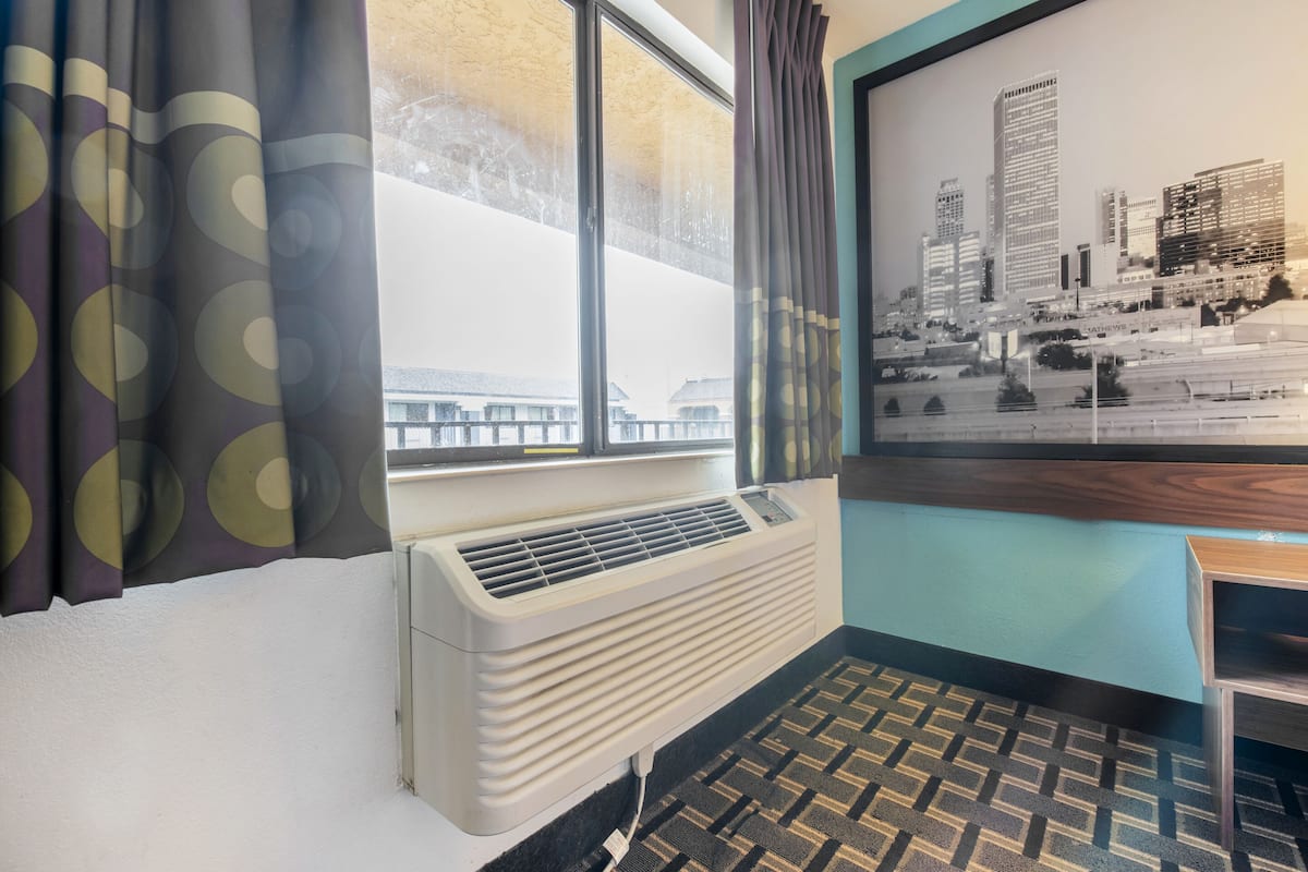 A room with a window, air conditioning unit, patterned carpet, and a framed cityscape picture on the wall.