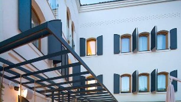 An outdoor patio area with a covered seating area, a table with chairs, and a building with arched windows in the background. - Sana Hotel Sarajevo (Sarajevo)