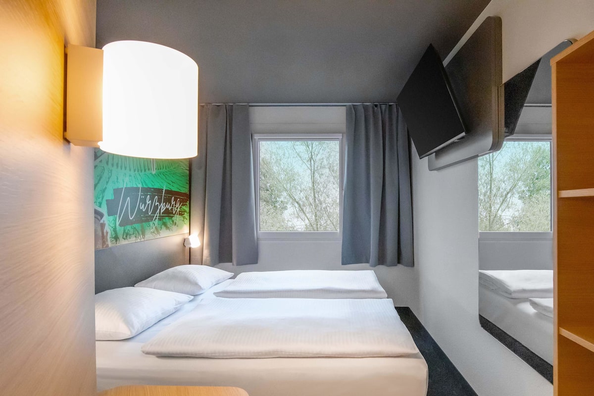 Une chambre d’hôtel avec deux lits, un téléviseur à écran plat fixé au mur, une grande fenêtre avec des rideaux et une lampe fixée au mur.