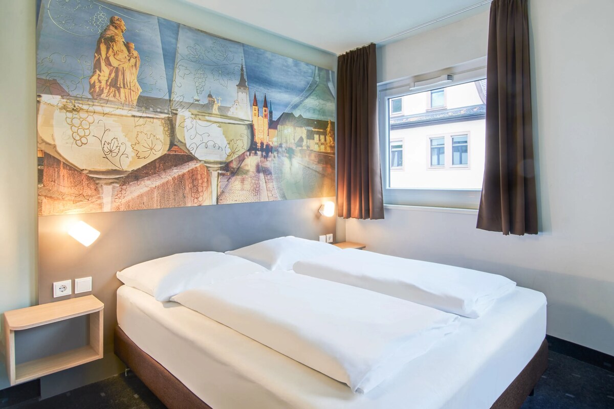 A hotel room with a large bed, a nightstand, a wall-mounted lamp, and a window with curtains.