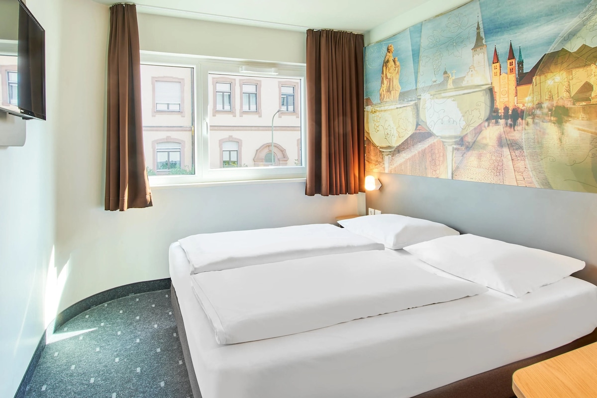 A hotel room with a bed, curtains, a painting of a cityscape, and a television.