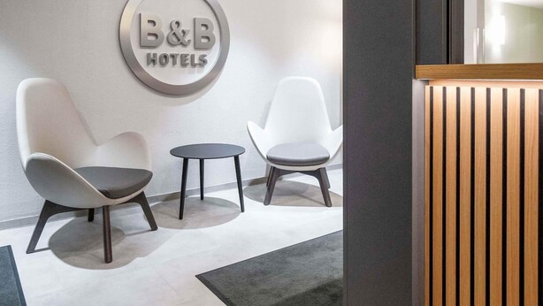 A hotel reception area with a circular sign reading "B&B HOTELS" on the wall, two modern chairs, a small round table, and a wooden reception desk. -  B&B HOTEL Würzburg-Ost (Würzburg)