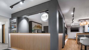 A modern hotel reception area with a wooden reception desk, a B&B sign, and a TV mounted on the wall. -  B&B HOTEL Würzburg-Ost (Würzburg)