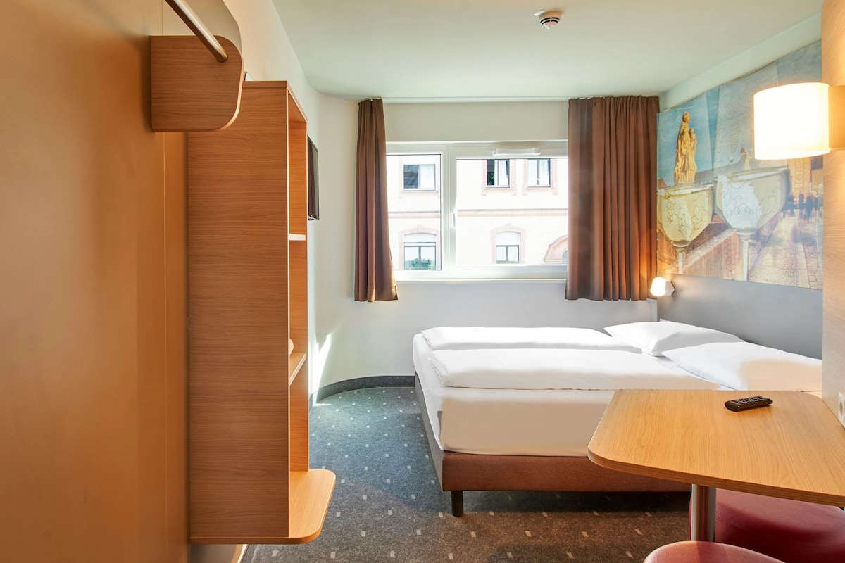 A hotel room with a bed, a desk, a chair, a window, and a painting.