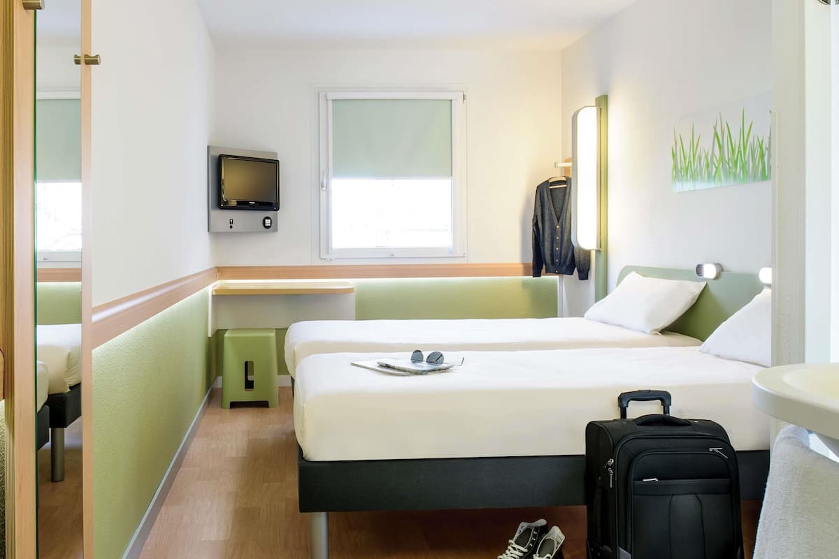 A hotel room with two beds, a TV, a window, a desk, and a suitcase.